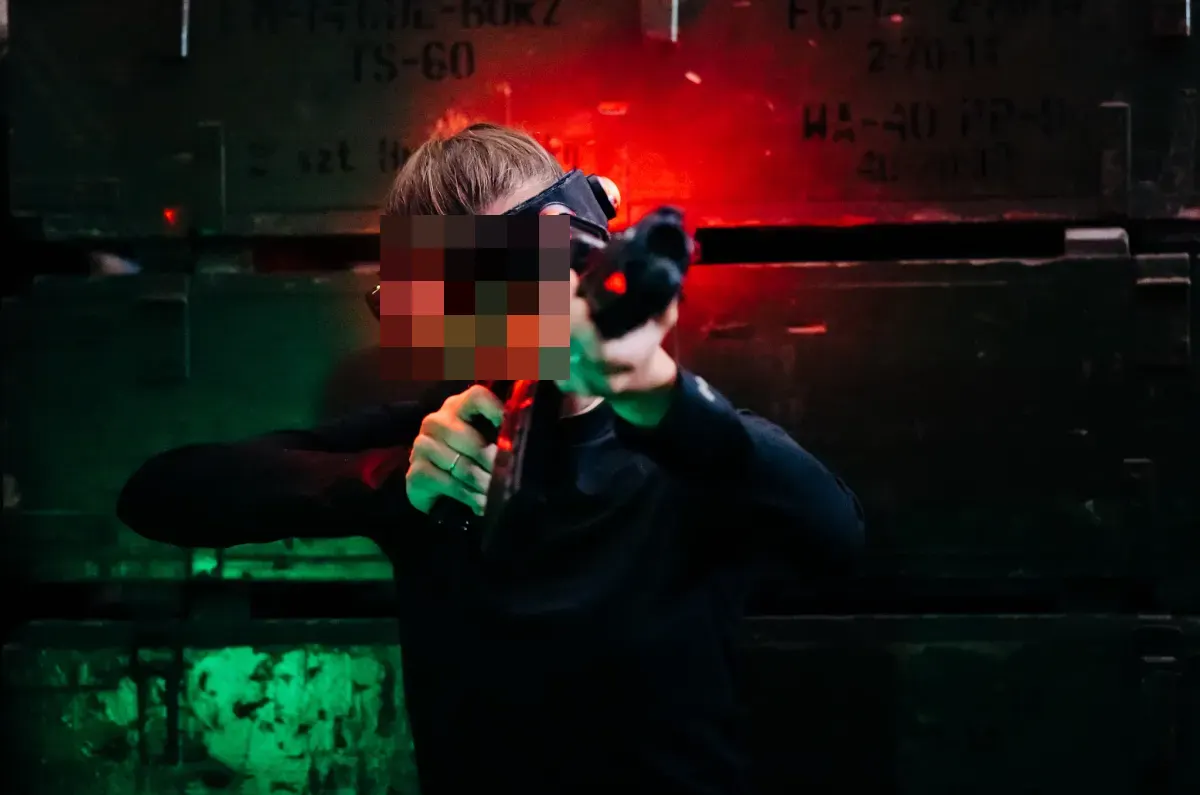 Gracz celuje na arenie, oświetlony czerwonym i zielonym światłem sprzętu, w tle drewniane skrzynie.
